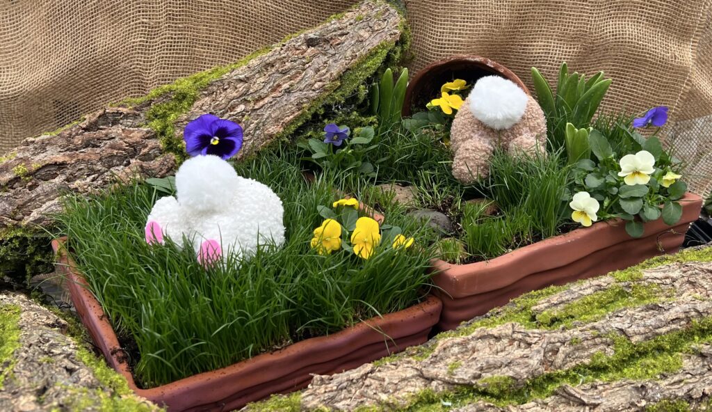 Two bunny decorations with fluffy tails appear to burrow into grass-filled planters surrounded by colorful flowers, bark, moss, and a broken pot, creating a playful spring scene.