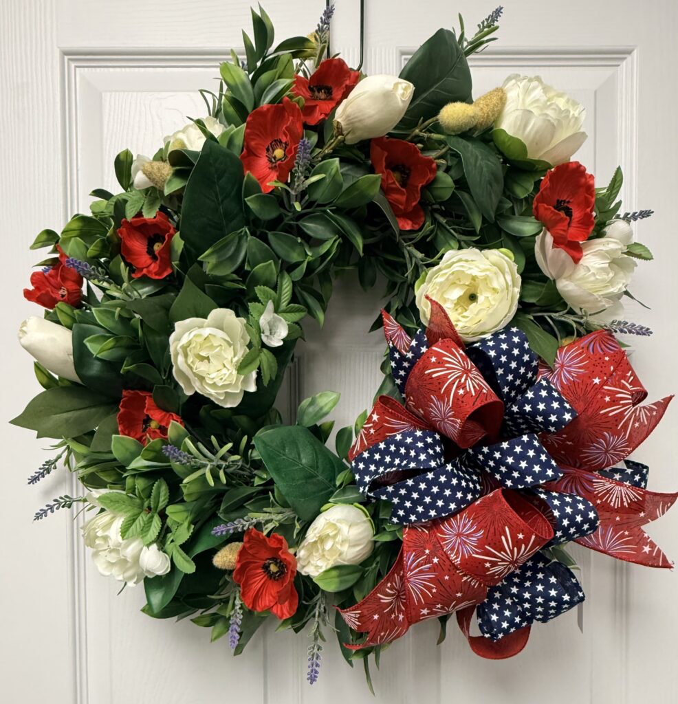 A festive wreath of green leaves, red poppies, white roses, and accent flowers, adorned with a large red, white, and blue star-patterned bow, hangs on a white paneled door.