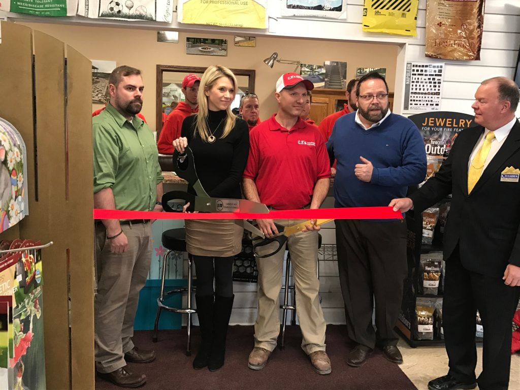 Kristin and Chad Parker at Timber Run Gardens opening stand indoors at a ribbon-cutting ceremony. One person in a red shirt holds oversized scissors cutting a red ribbon, while others smile nearby. Shelves and products are visible in the background.