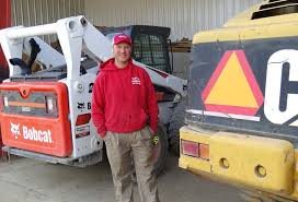 A man in a red hoodie and cap stands between a white Bobcat loader and a yellow machine with a reflective safety triangle, inside a garage or workshop area.