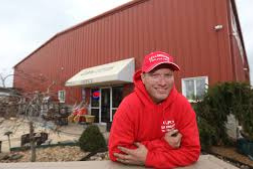 Chad Parker in a red hoodie and red cap smiles while leaning on a surface in front of a large red building with a tan awning and a visible “OPEN” sign by the entrance.