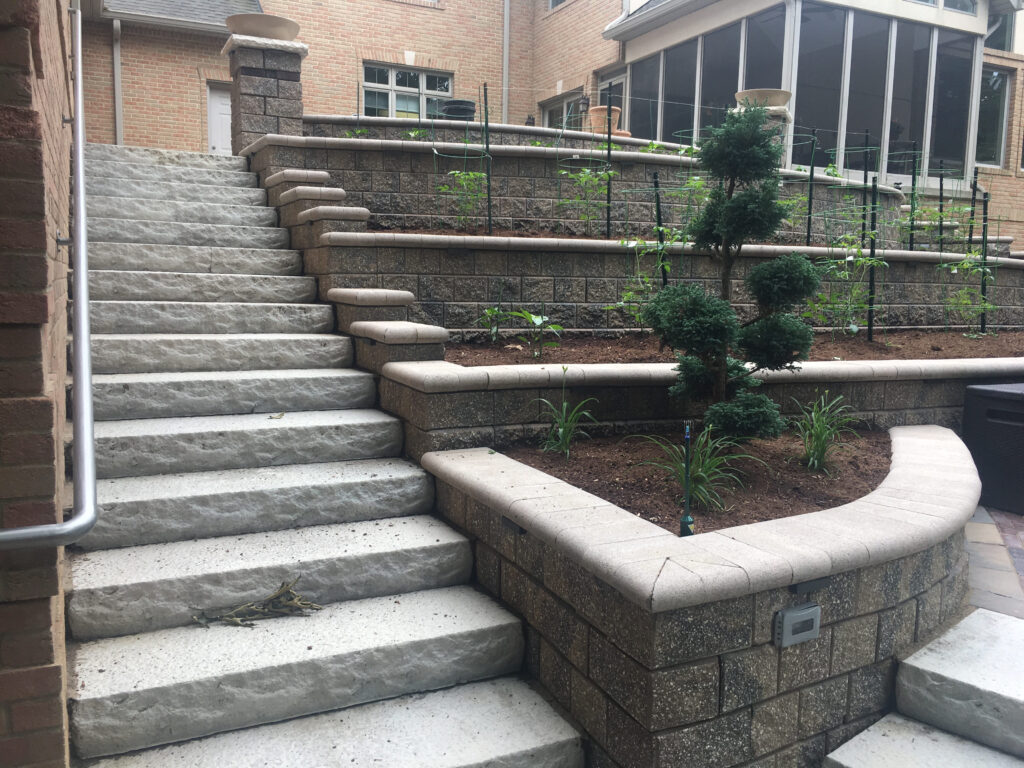 Wide concrete steps ascend beside tiered retaining walls with garden beds containing small plants and shrubs. The walls and steps are adjacent to a brick house with a screened porch above.