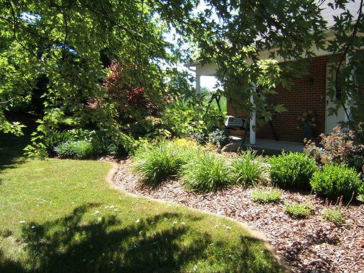 A shaded yard with a mulched garden bed, green shrubs, flowering plants, and a grassy lawn beside a brick house with a covered porch and a porch swing. Sunlight filters through the leafy trees overhead.