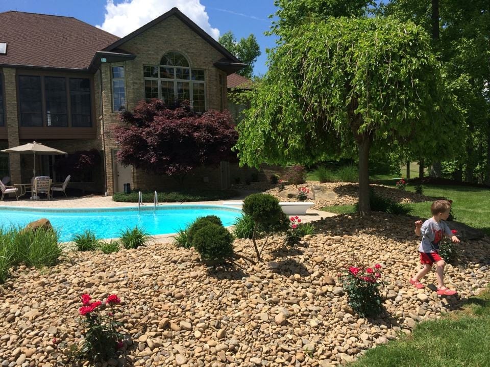 A young child in red shorts runs on a rocky area near a backyard pool, surrounded by plants and flowers, with a large brick house and trees in the background on a sunny day.