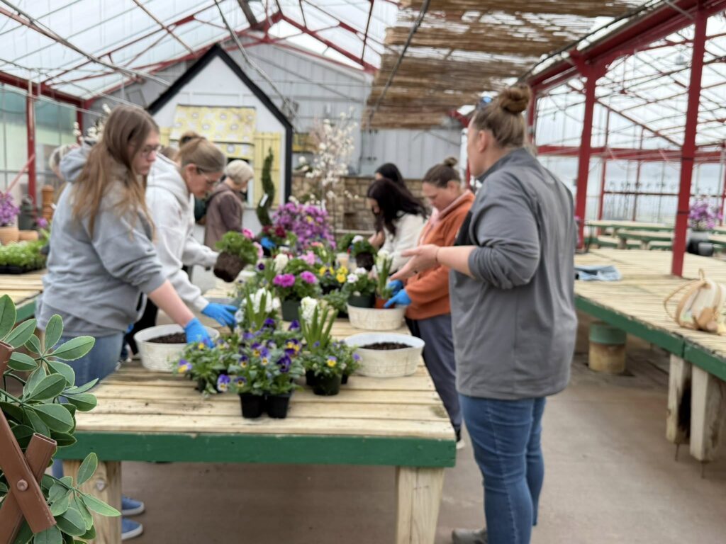 A group of women work together at a long table in a greenhouse, arranging colorful flowers and plants in pots. The atmosphere appears collaborative and creative.