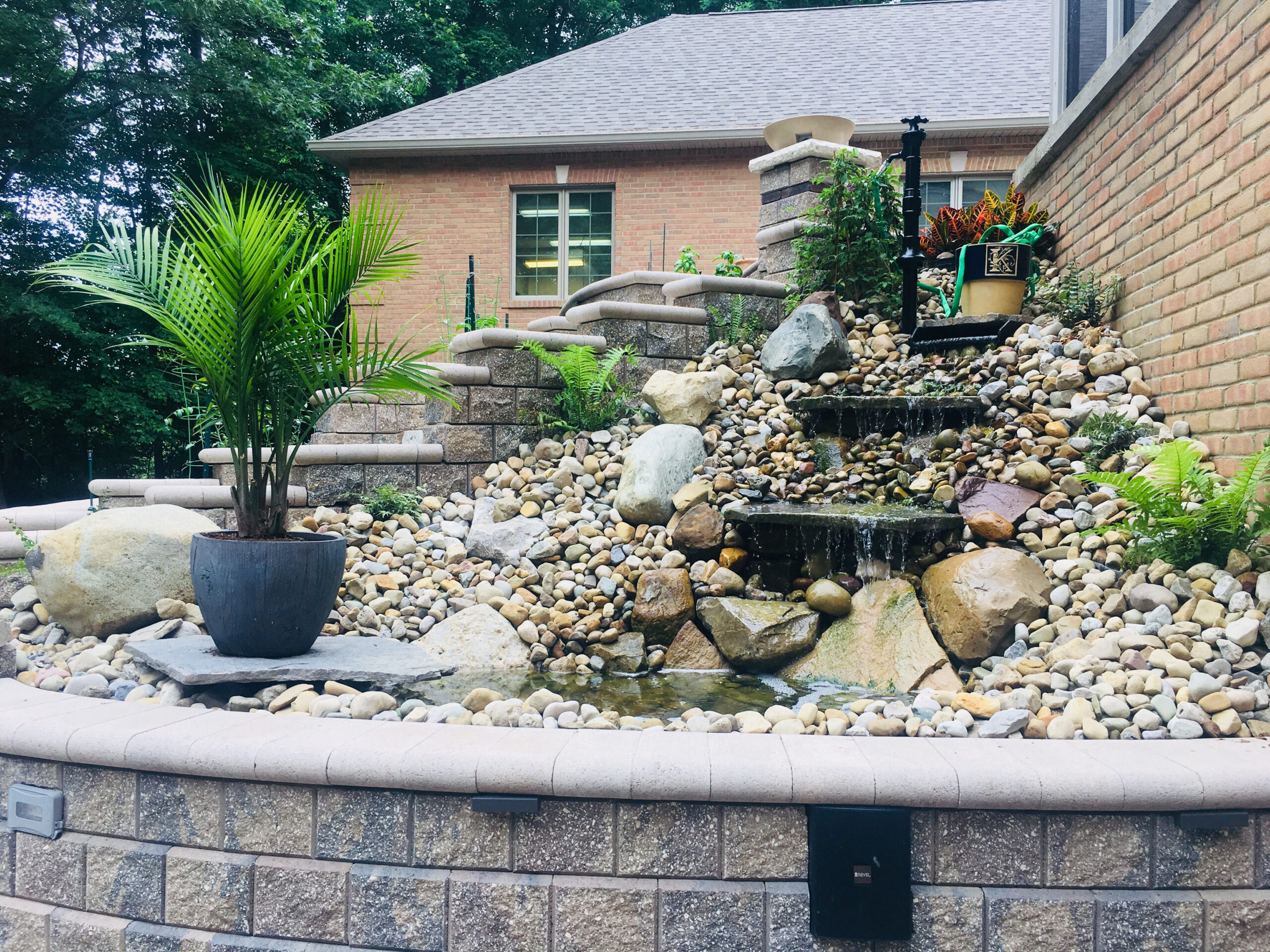 A landscaped backyard water feature with a small waterfall flows over rocks, surrounded by ferns, a potted palm, and other plants, with a brick house and stone retaining wall in the background.
