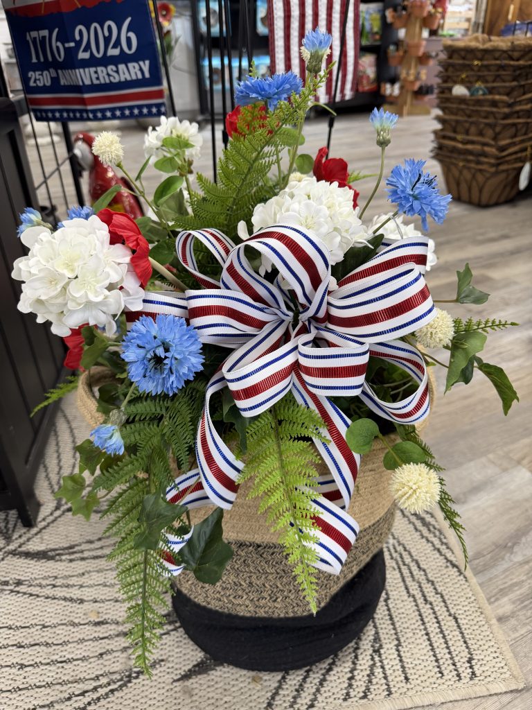 A floral arrangement of red, white, and blue flowers in a woven basket, accented with a patriotic striped ribbon bow.