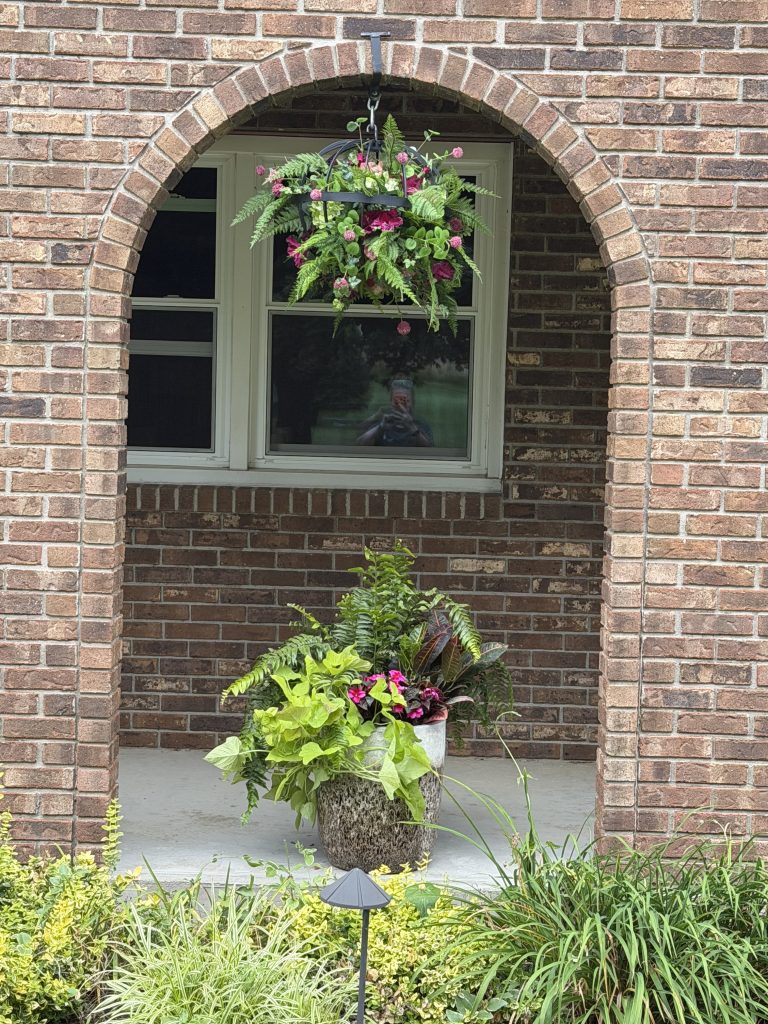 A brick arched entryway at a plant nursery featuring a hanging basket of pink and green flowers above a large floor planter filled with ferns and foliage.